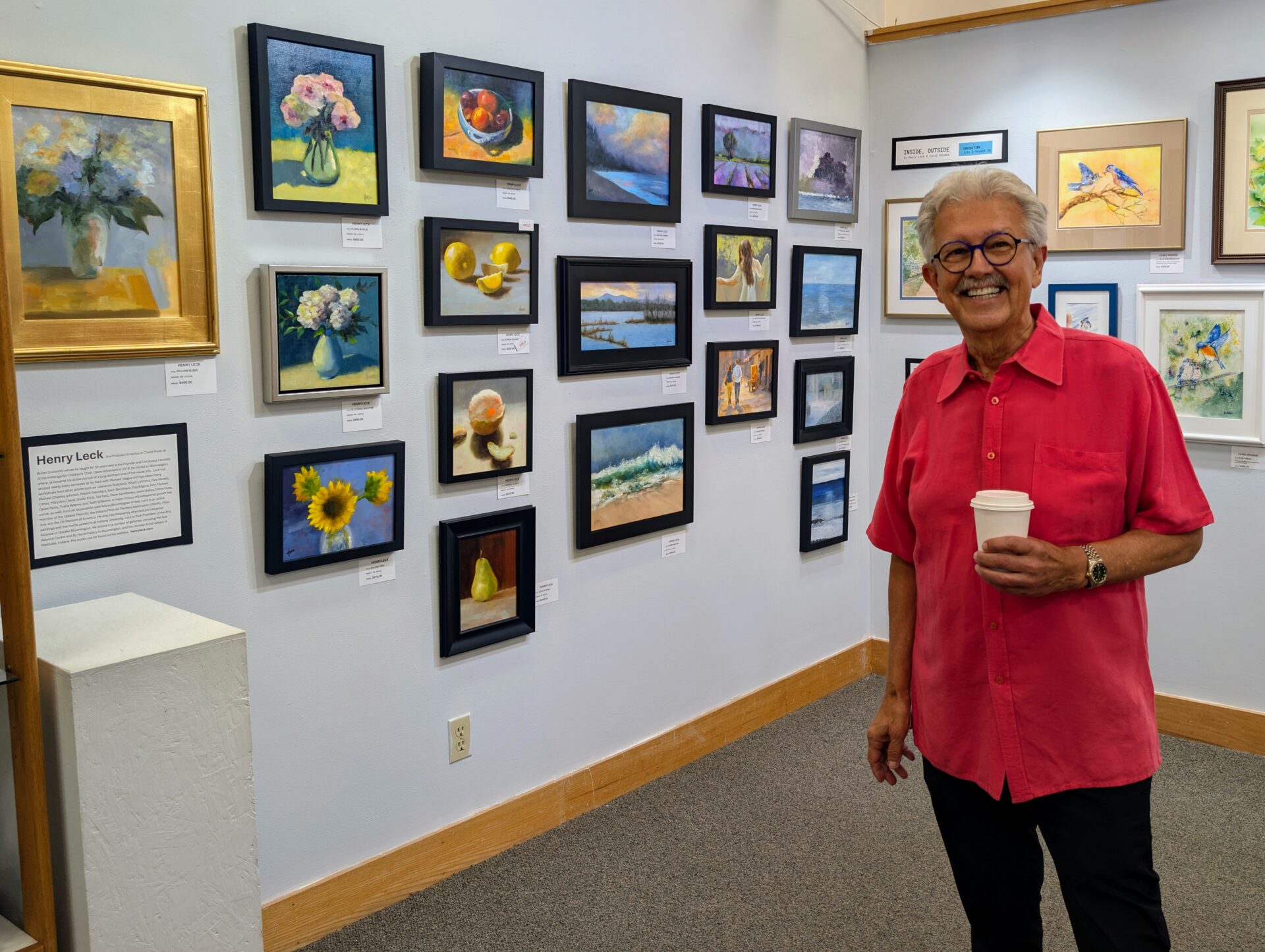Artist Henry Leck smiling beside a wall of framed paintings at his By Hand Gallery exhibition reception